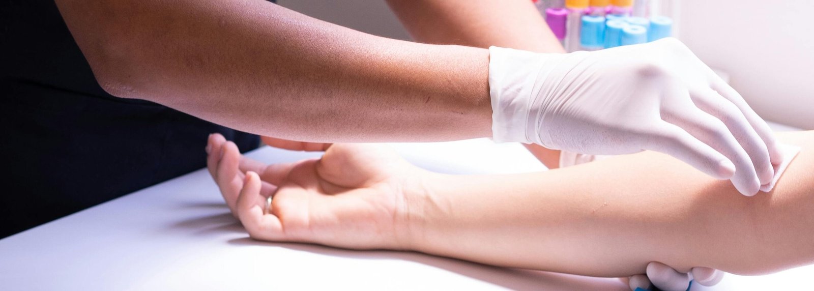 Healthcare worker in black scrubs preparing to draw blood from a patient's arm using gloves and medical equipment.