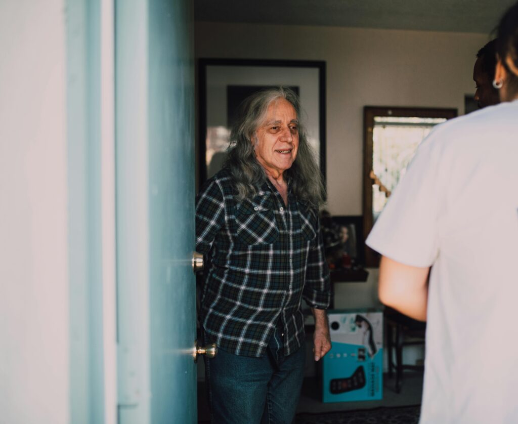 Older man with long grey hair opening his front door to a community health worker during a home visit.